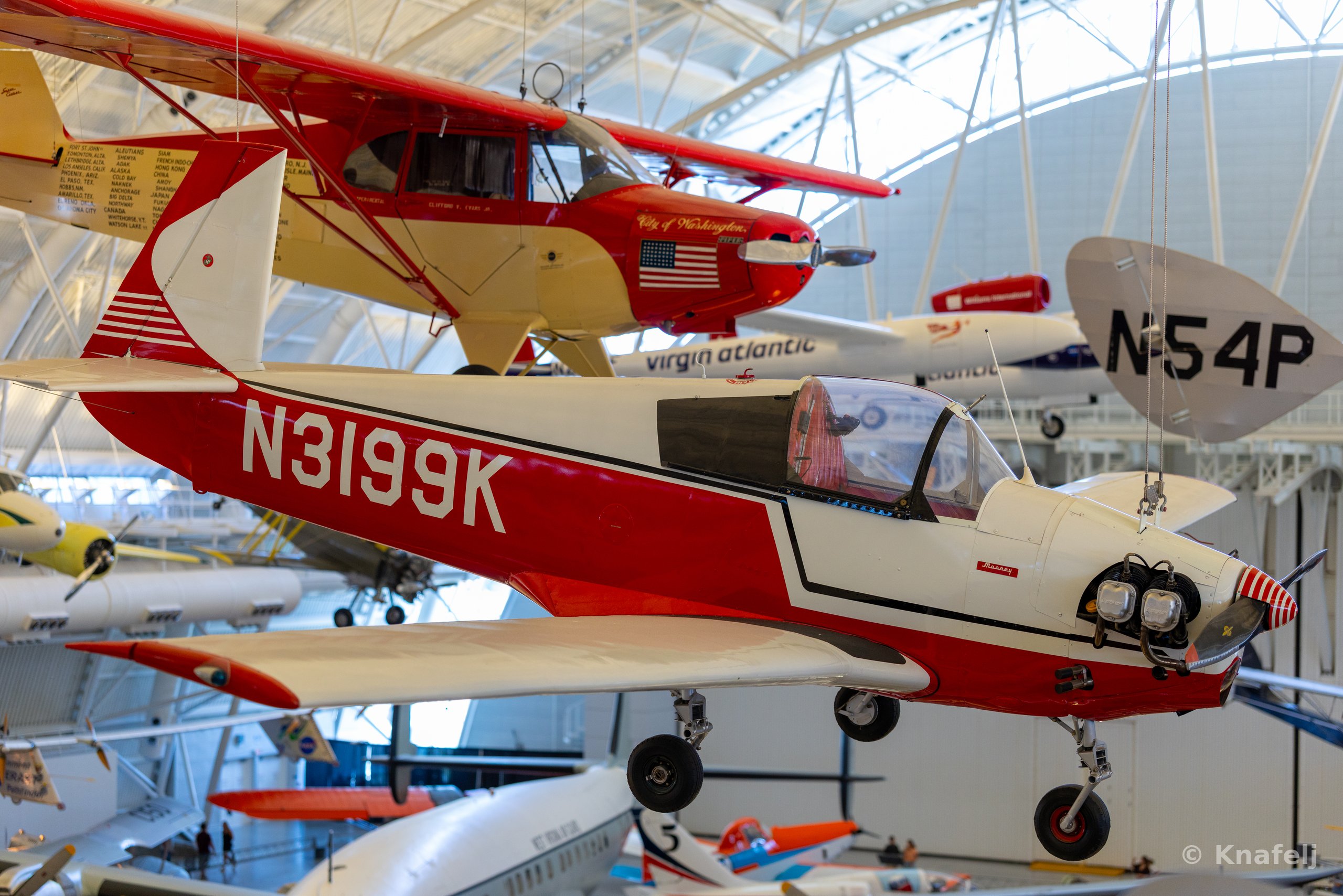Multiple small vintage airplanes suspended from the ceiling inside a large museum hangar, with a prominent red and white aircraft labeled N3199K in the foreground and other planes marked N54P and City of Washington above and behind it.