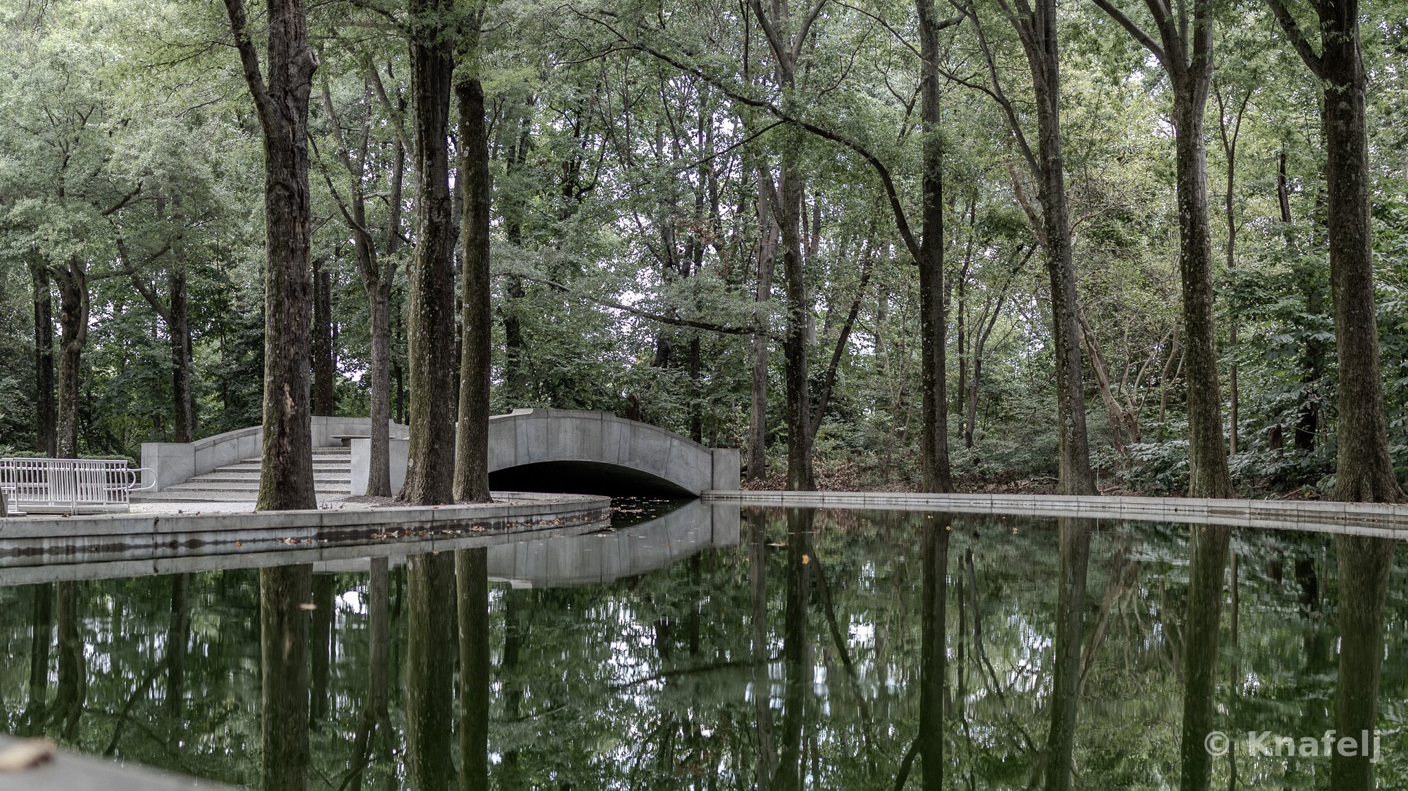 A gray stone arched bridge crossing a still body of water in a wooded park, surrounded by tall trees that are reflected in the calm surface, with a metal railing and steps on the left bank.