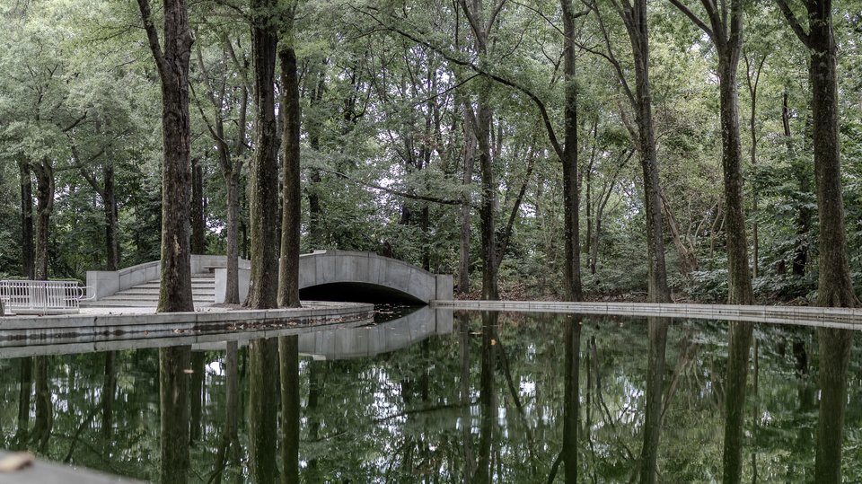 A gray stone arched bridge crossing a still body of water in a wooded park, surrounded by tall trees that are reflected in the calm surface, with a metal railing and steps on the left bank.