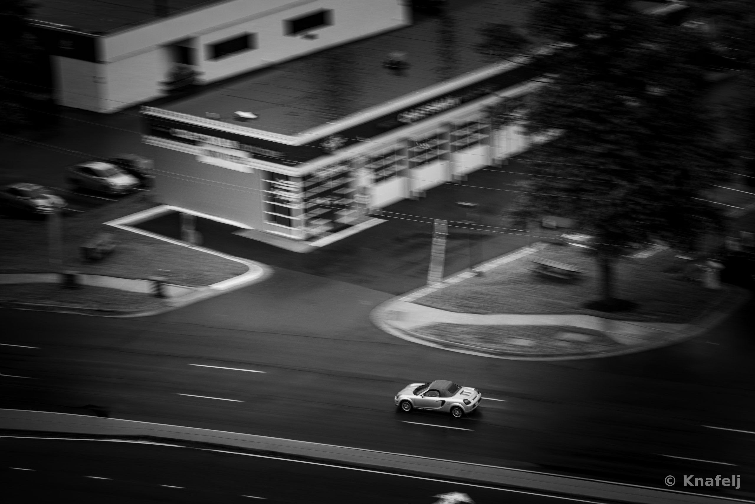 Black and white aerial view of a silver convertible sports car driving on a road, with motion blur streaking a building, trees, and parking lot in the background.