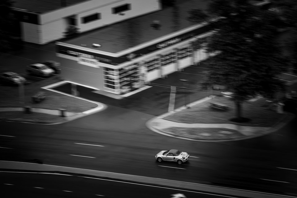Black and white aerial view of a silver convertible sports car driving on a road, with motion blur streaking a building, trees, and parking lot in the background.