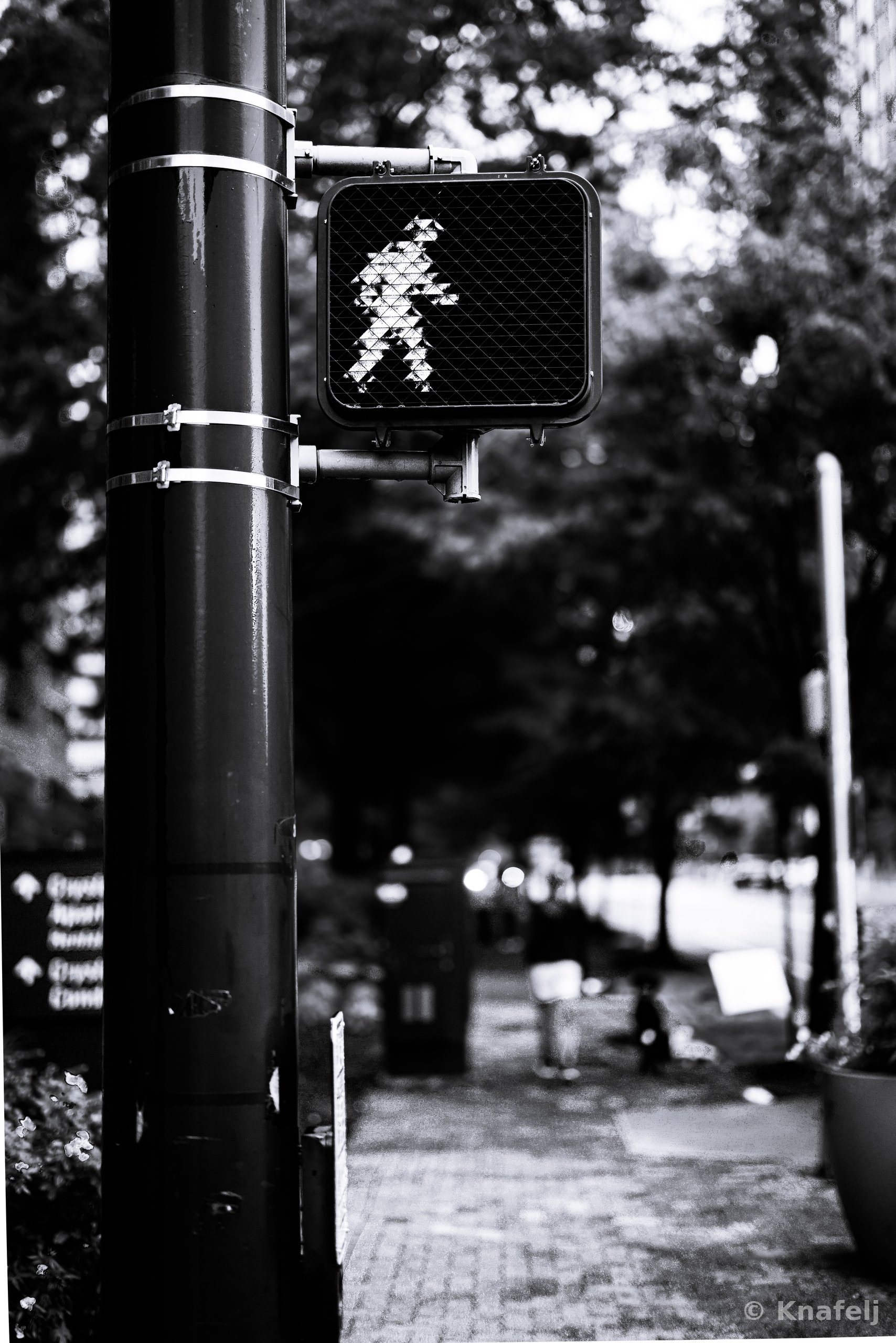 Black and white photo of a pedestrian crossing signal showing a walking figure, mounted on a large black pole, with a brick sidewalk, trees, and blurred pedestrians in the background.