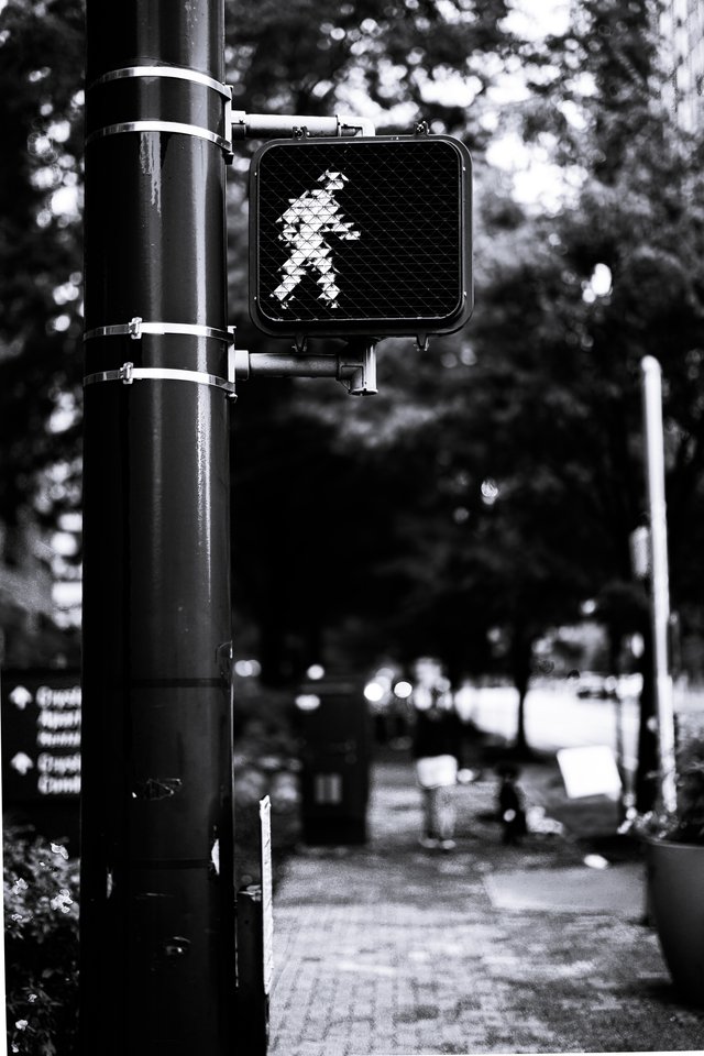 Black and white photo of a pedestrian crossing signal showing a walking figure, mounted on a large black pole, with a brick sidewalk, trees, and blurred pedestrians in the background.