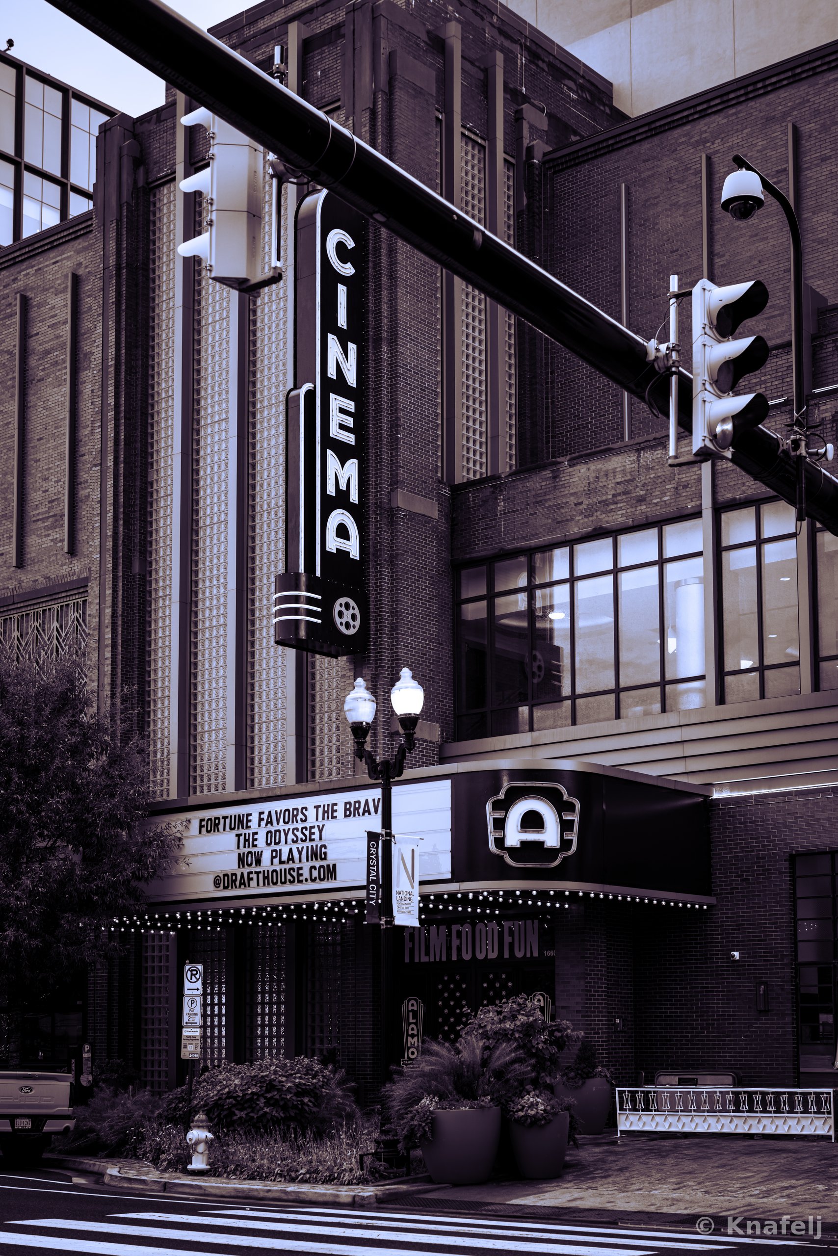 A tall vertical 'CINEMA' sign mounted on a brick art deco movie theater, with an illuminated marquee reading 'Fortune Favors the Brave, The Odyssey, Now Playing' below. A traffic light and ornate lamppost are in the foreground, toned in monochrome purple.