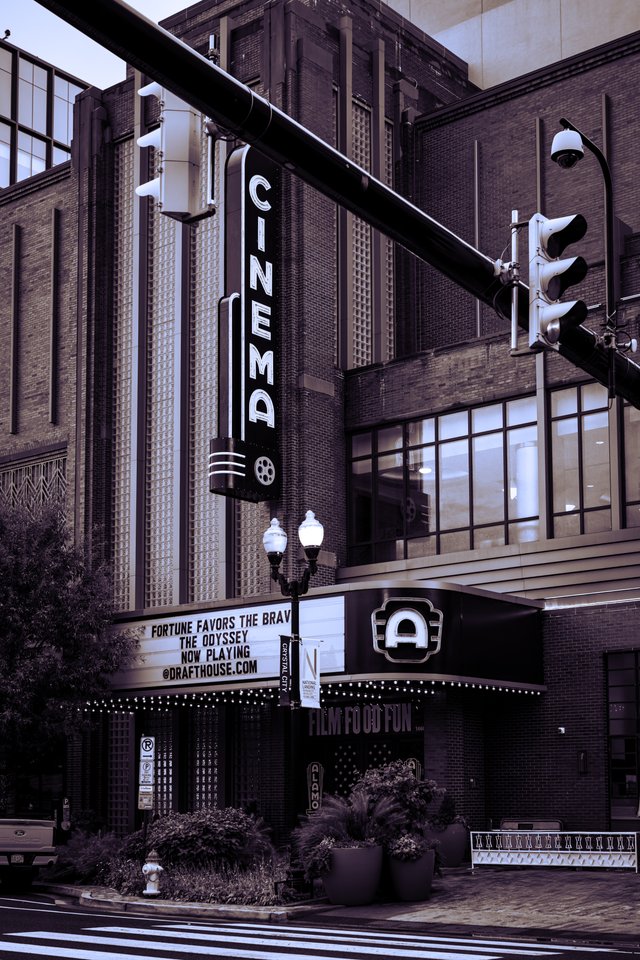 A tall vertical 'CINEMA' sign mounted on a brick art deco movie theater, with an illuminated marquee reading 'Fortune Favors the Brave, The Odyssey, Now Playing' below. A traffic light and ornate lamppost are in the foreground, toned in monochrome purple.