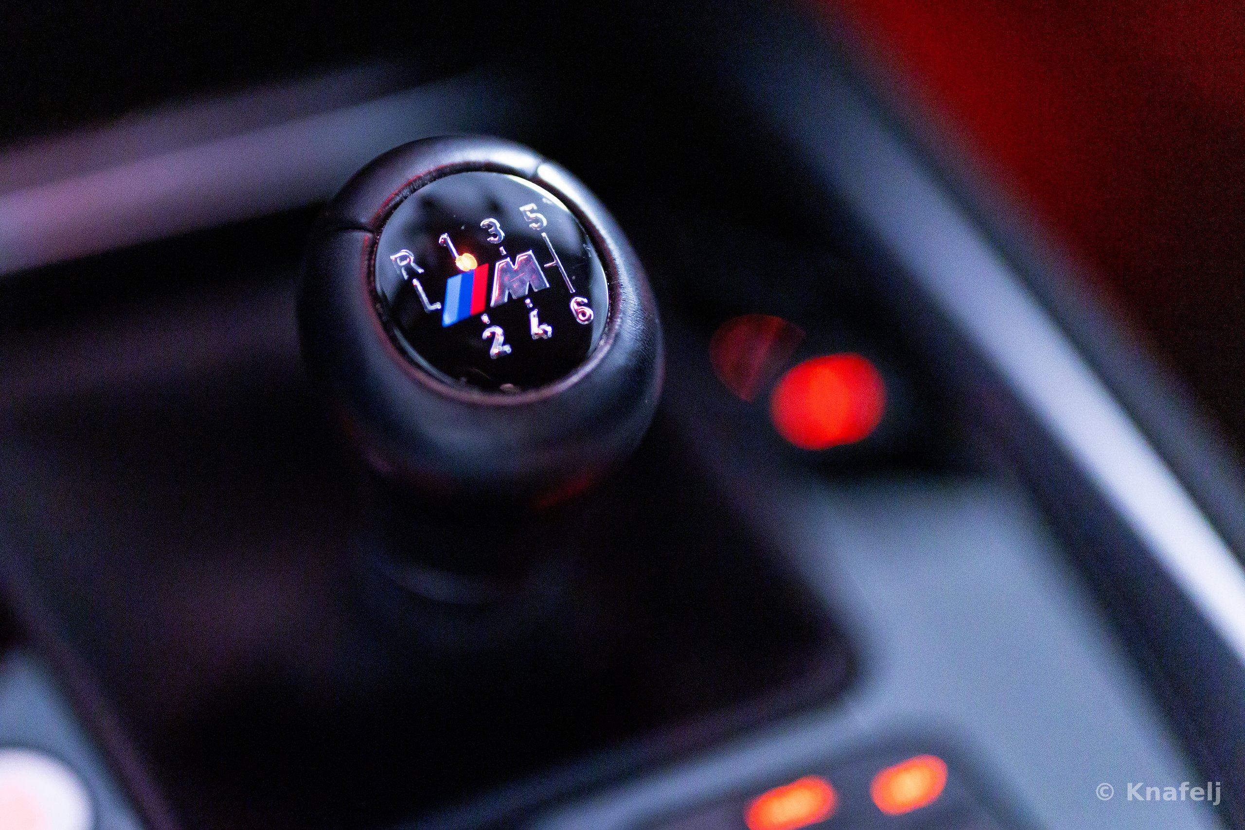 Black leather BMW M manual gear shift knob showing the shift pattern with reverse and gears one through six, and the blue, purple, and red M logo, photographed close-up with blurred red lights in the background.