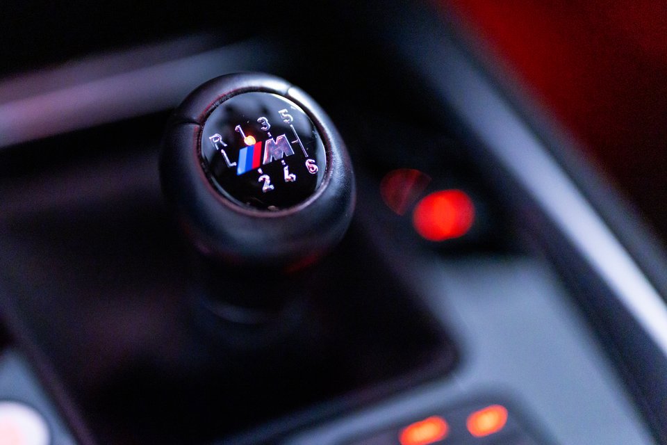 Black leather BMW M manual gear shift knob showing the shift pattern with reverse and gears one through six, and the blue, purple, and red M logo, photographed close-up with blurred red lights in the background.