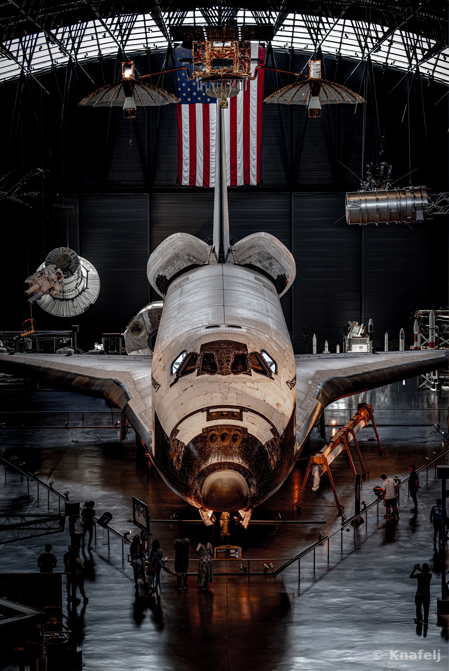 A retired space shuttle displayed nose-first inside a large museum hangar, with an American flag hanging above it and small groups of people walking around its base.