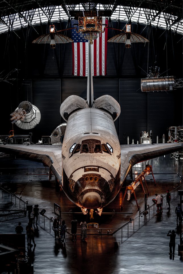 A retired space shuttle displayed nose-first inside a large museum hangar, with an American flag hanging above it and small groups of people walking around its base.