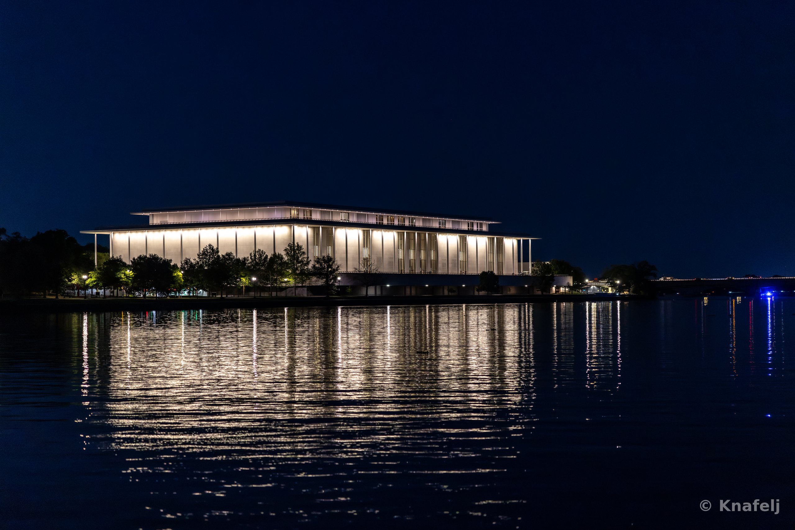 A large brightly lit modern building with tall columns sits along a river at night, its warm lights reflecting on the dark rippling water beneath a deep blue sky.