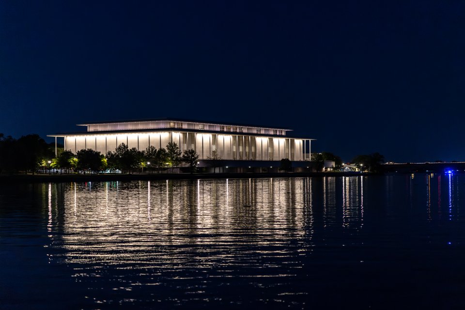 A large brightly lit modern building with tall columns sits along a river at night, its warm lights reflecting on the dark rippling water beneath a deep blue sky.