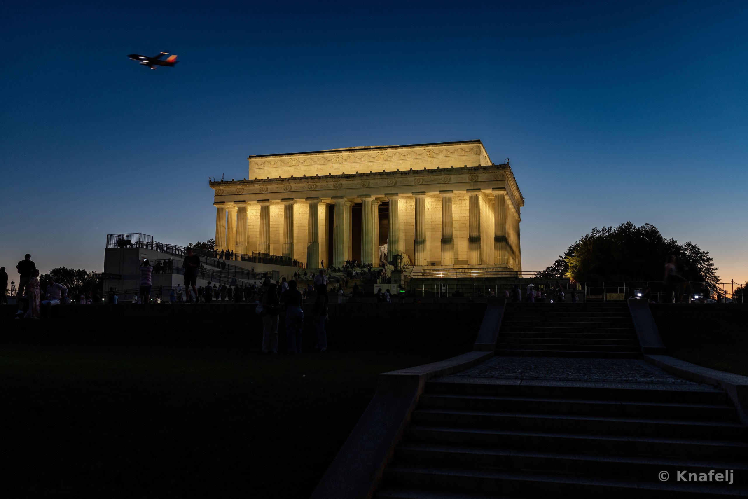 The illuminated Lincoln Memorial at dusk with columns lit by warm light, silhouetted people on the steps and foreground stairs, and a small plane flying in the darkening blue sky.