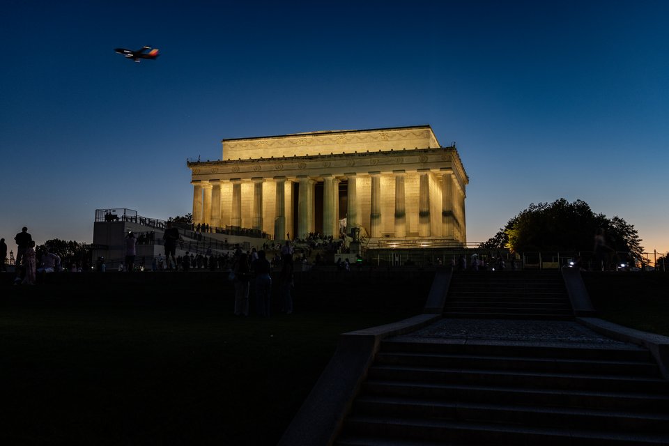 The illuminated Lincoln Memorial at dusk with columns lit by warm light, silhouetted people on the steps and foreground stairs, and a small plane flying in the darkening blue sky.