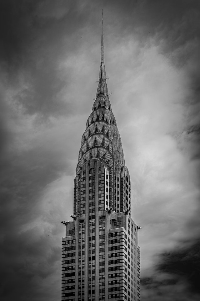 Black and white photograph of the upper portion of the Chrysler Building, showing its terraced Art Deco crown, radiating steel arches, and tall spire against a moody, cloudy sky.